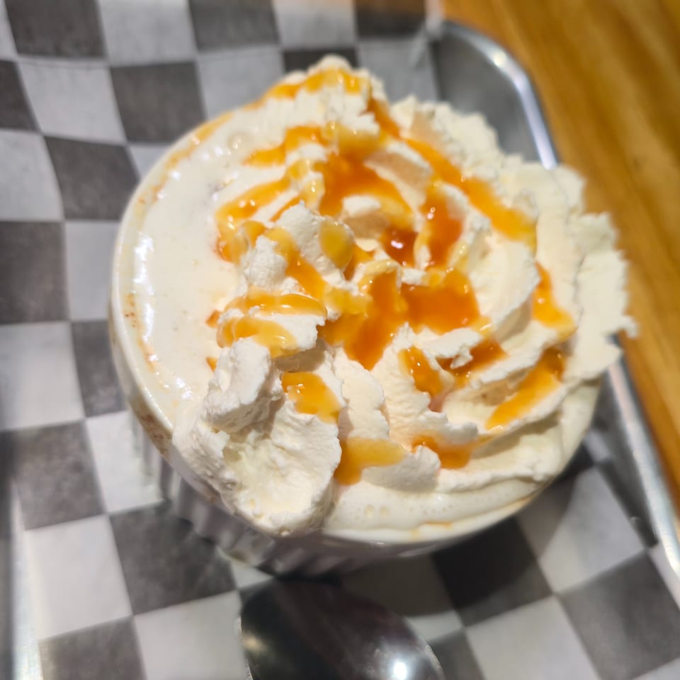 A white mug of coffee topped with whipped cream and caramel drizzle on a checkered paper.