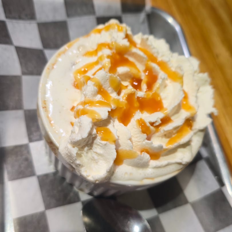 A white mug of coffee topped with whipped cream and caramel drizzle on a checkered paper.
