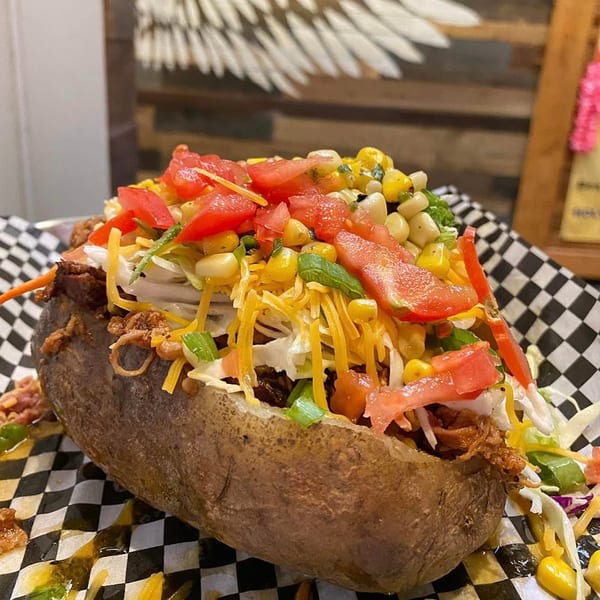 Holy Smokes' loaded baked potato overflowing with pulled pork, cheese, corn, and tomatoes on checkered paper.