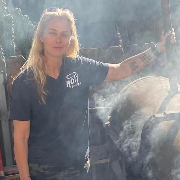 A woman in a Holy Smokes t-shirt stands next to a smoking barbecue pit.